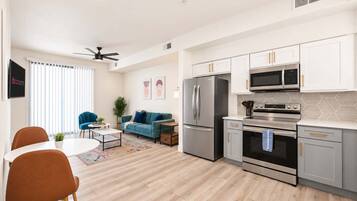 A modern kitchen with stainless steel appliances, a dining area with a white table and orange chairs, and a living room with a blue sofa.