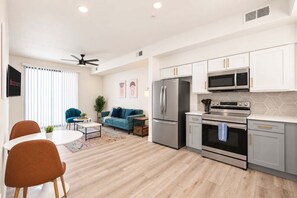 A modern kitchen with stainless steel appliances, a dining area with a white table and orange chairs, and a living room with a blue sofa. - CozySuites Glendale - Westgate w pool! 1BR 1BA (Glendale)