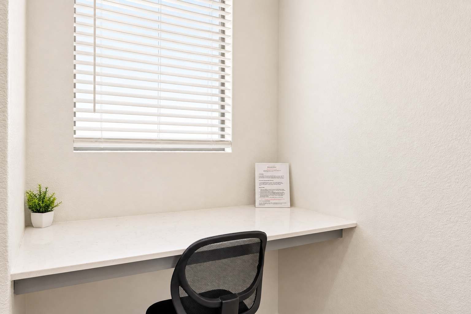 A small office space with a white desk, a black mesh chair, a potted plant, and a window with blinds.