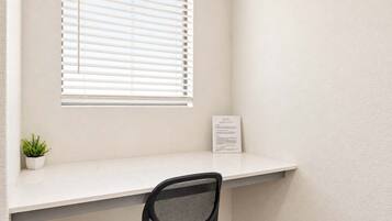 A small office space with a white desk, a black mesh chair, a potted plant, and a window with blinds.