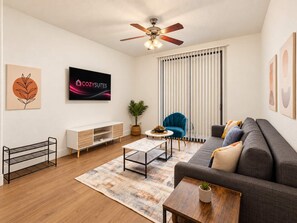 A modern living room with a sofa, a coffee table, a TV, a ceiling fan, and a window with blinds. - CozySuites Glendale - Westgate w pool! 2BR 2BA (Glendale)