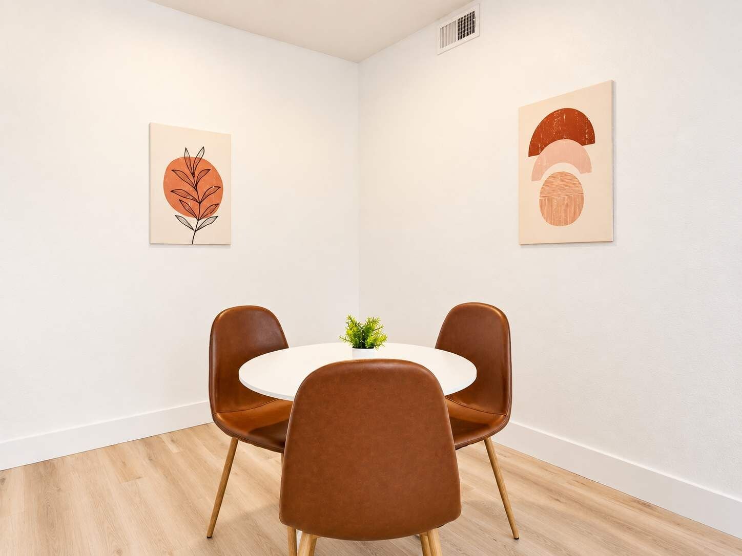 A room with a round table, two brown chairs, a small plant, and two wall-mounted artworks.