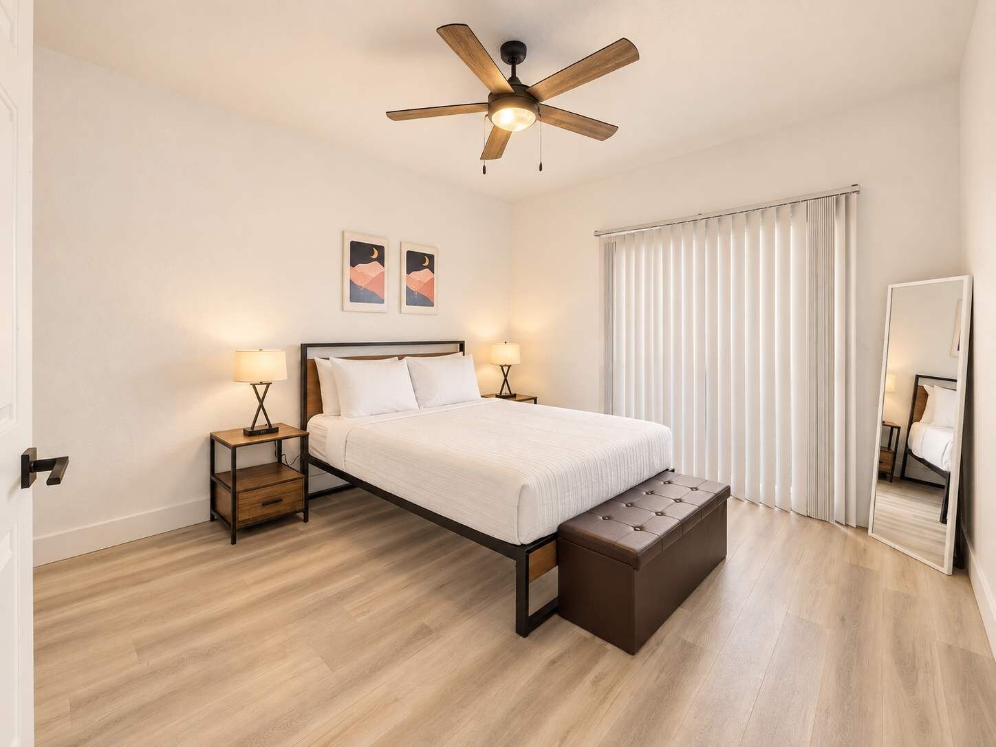Ein Hotelzimmer mit Bett, Nachttischlampen, Deckenventilator, großem Fenster mit Jalousien und einem Spiegel.