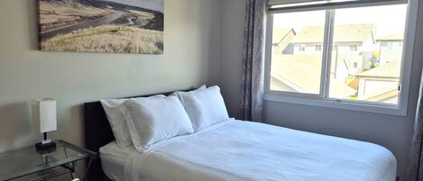A hotel room with a bed, a desk with a lamp, a chair, a window with curtains, and a painting on the wall.