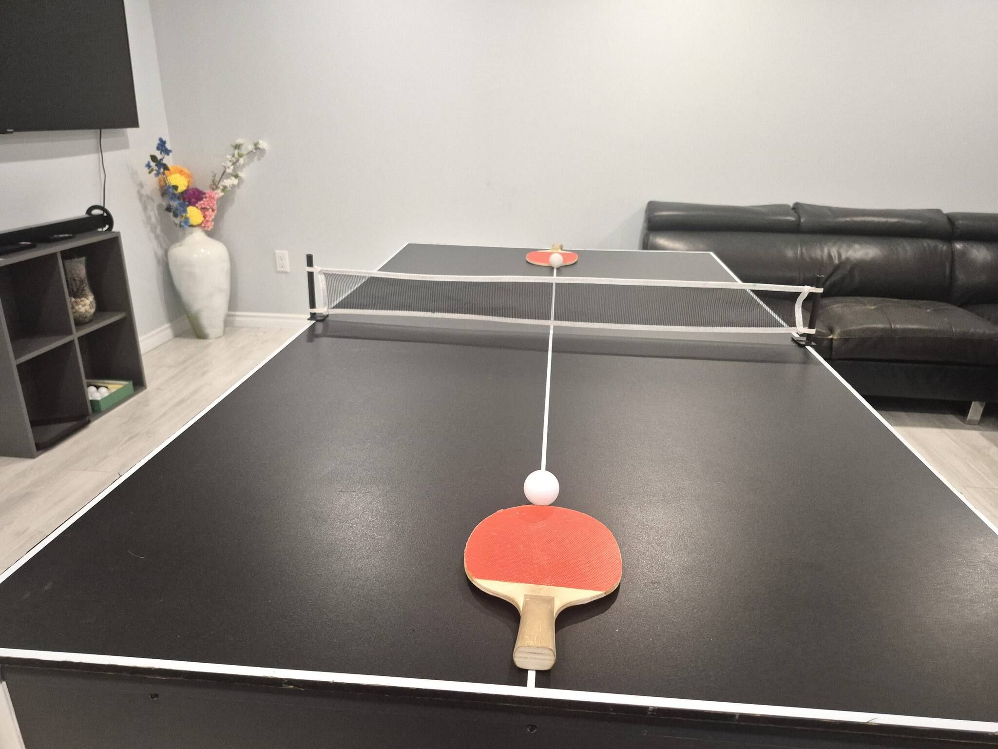 Une table de ping-pong avec une balle et une raquette au premier plan, un canapé en cuir noir et un vase de fleurs à l’arrière-plan.