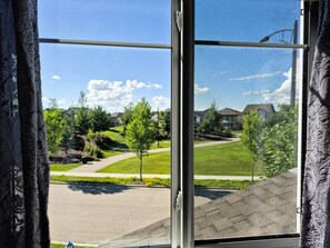 Blick durch ein Fenster mit Metallrahmen und einem Moskitonetz auf ein Wohngebiet mit Häusern, Bäumen und blauem Himmel.
