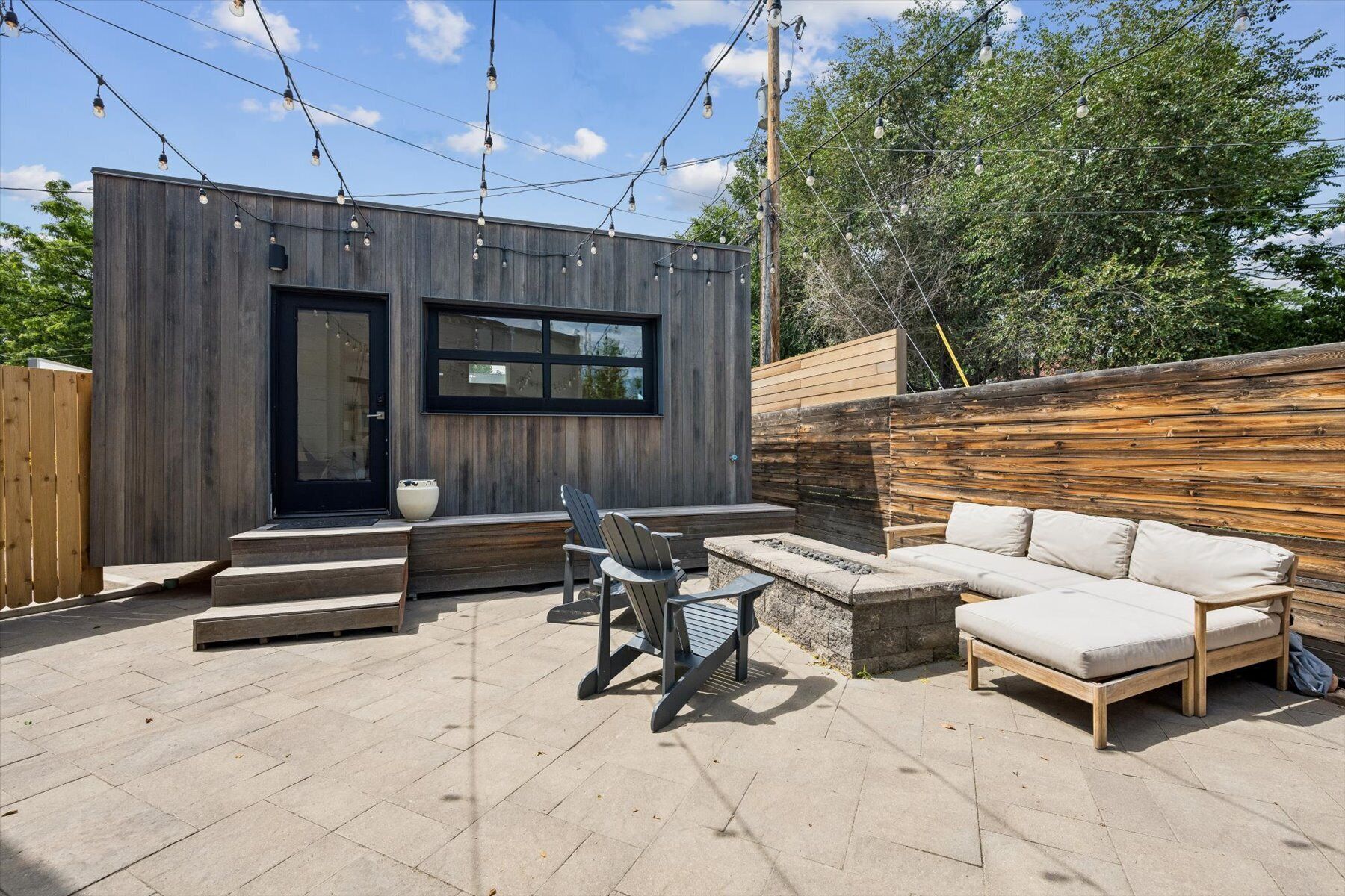 Une terrasse extérieure moderne, dotée d’une plateforme en bois, d’une porte noire, d’un espace salon avec un canapé et des fauteuils, et de guirlandes lumineuses.