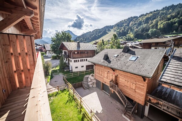 A wooden chalet with a stone staircase, a view of a village, and a mountain in the background. - ARKOS 202: Beautiful two-bedroom apartment for 4–5 guests in the heart of Morzine (MORZINE)