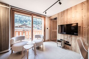 A modern living room with a large TV, two sofas, a small table, and a balcony with a view of a mountain village. - ARKOS 202: Beautiful two-bedroom apartment for 4–5 guests in the heart of Morzine (MORZINE)