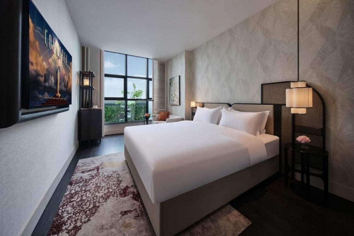 A modern hotel room with a large bed, a view of greenery outside, and a framed picture on the wall.