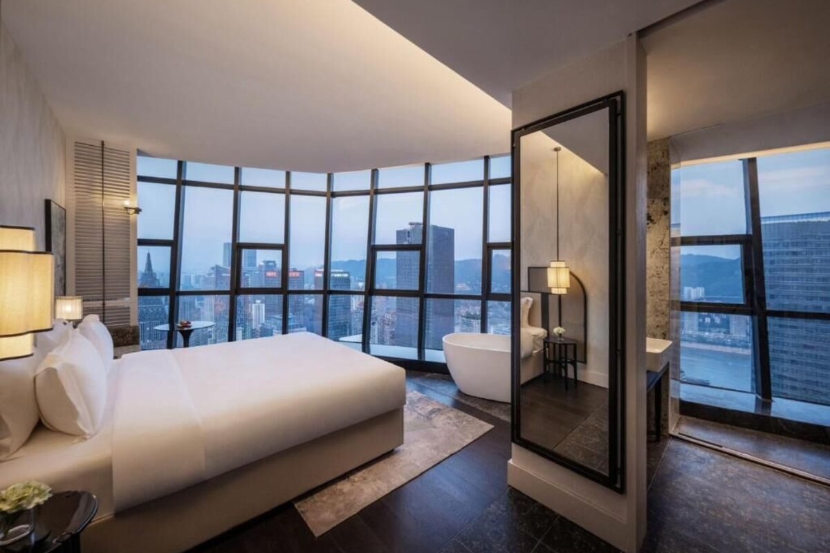 A modern hotel room with a large bed, a bathtub, and a city view through floor-to-ceiling windows.
