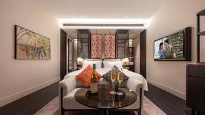 Quarto de hotel com cama, mesa com copos e garrafa, e obras de arte nas paredes.
