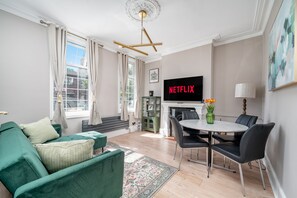A living room with a green sofa, a dining table with chairs, a flat-screen TV, and a fireplace. - Fulham s Best-kept Secret 2 Bed Walled Garden (London)
