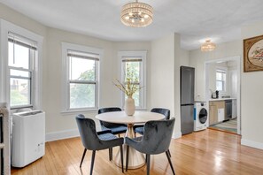 A modern kitchen with a built-in washer and dryer, a dining area with a round table and chairs, and large windows providing natural light. - Sunny & Spacious 2BR Apartment Near Harvard | Long Stays (Watertown)