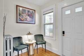 A room with a white door, a framed painting, two chairs, a small table, and a lamp. - Sunny & Spacious 2BR Apartment Near Harvard | Long Stays (Watertown)
