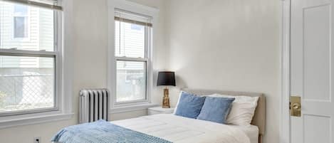 Un dormitorio con cama, dos ventanas, un radiador, una lámpara de noche y una puerta.
