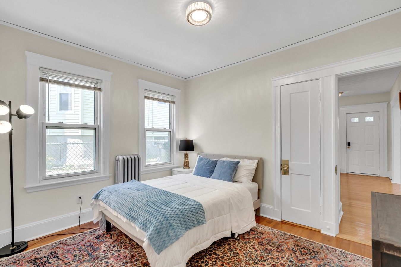 A bedroom with a bed, two windows, a radiator, a lamp, and a patterned rug.