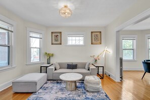 A modern living room with a grey sofa, a round coffee table, a side table, and a floor lamp. There are two windows with blinds, a chandelier, and framed artwork on the wall. - Sunny & Spacious 2BR Apartment Near Harvard | Long Stays (Watertown)