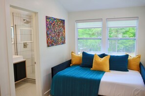 A bedroom with a bed, a window with a view of trees, and a bathroom with a tiled wall and a shower. - Cozy Mid-Century Modern Hideaway (Durham)
