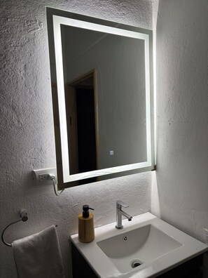 Un baño con un lavamanos blanco, un espejo con luz y una toalla colgada en un perchero.