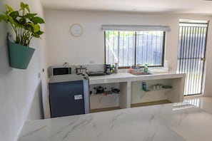 Cocina compacta con refrigerador, estufa y microondas. Hay una ventana con persianas, un reloj en la pared y una planta en maceta.