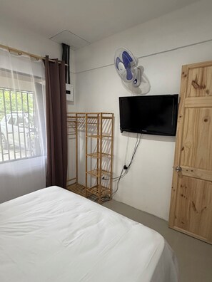 Habitación de hotel con cama, armario de madera, televisor montado en la pared, ventilador y ventana con cortinas.