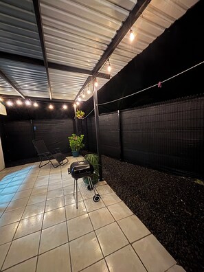 A covered patio area with a grill, lounge chairs, and string lights.