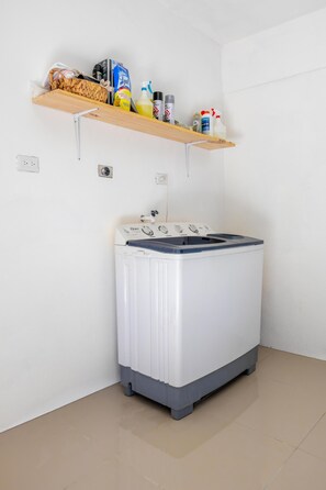 A laundry room with a front-loading washing machine, a shelf with cleaning supplies, and a power outlet.
