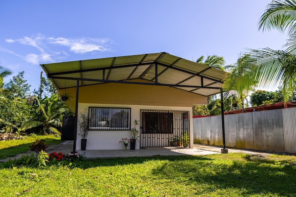 Una casa de un piso con un porche cubierto, patio cercado y follaje tropical.