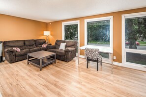 Ein Wohnzimmer mit braunen Sofas, einem Couchtisch, einem gemusterten Sessel und großen Fenstern mit Blick auf Bäume und eine Straße.