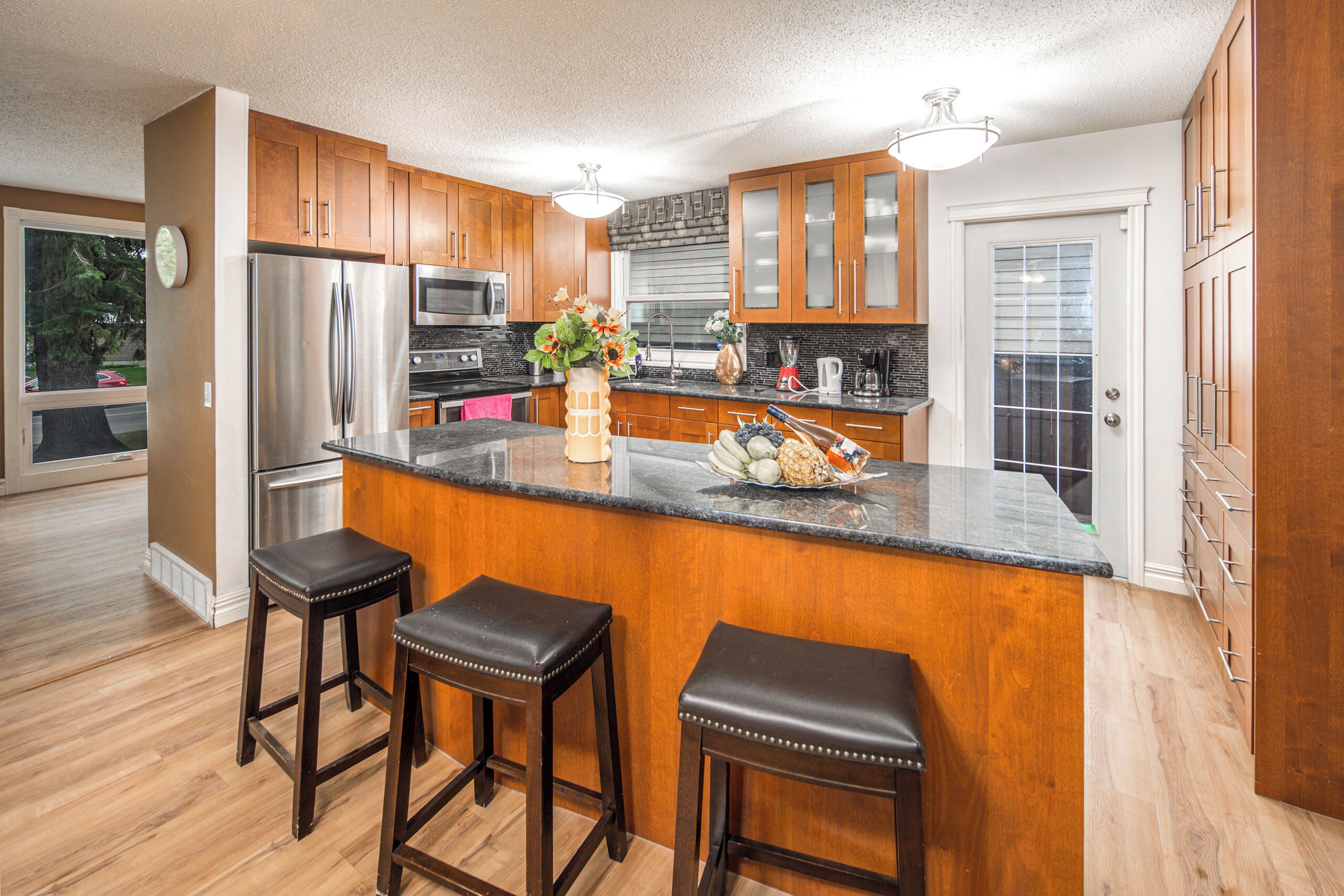 A modern kitchen with wooden cabinets, stainless steel appliances, a marble countertop, and two bar stools.