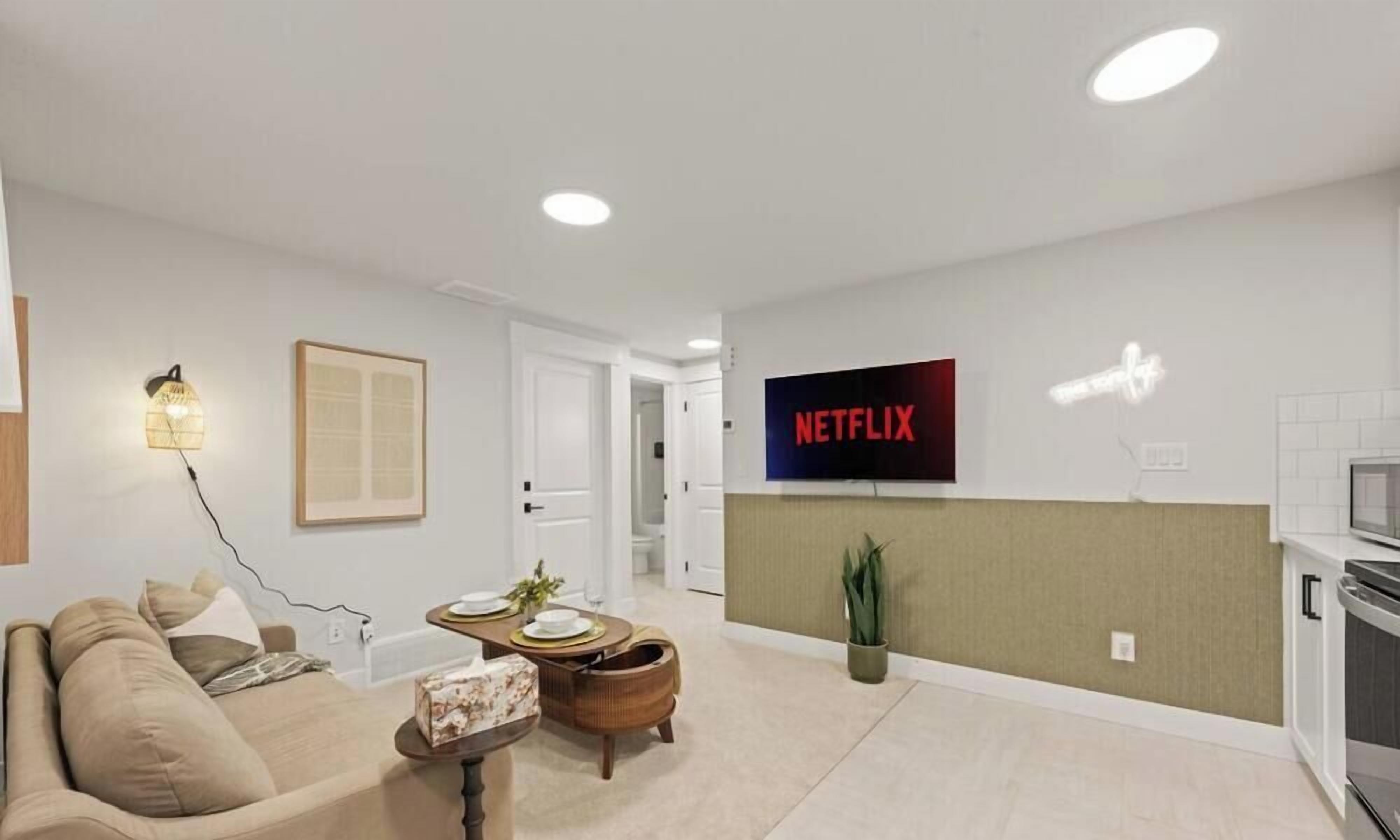A modern living room with a sofa, a dining table, and a wall-mounted television displaying the Netflix logo.