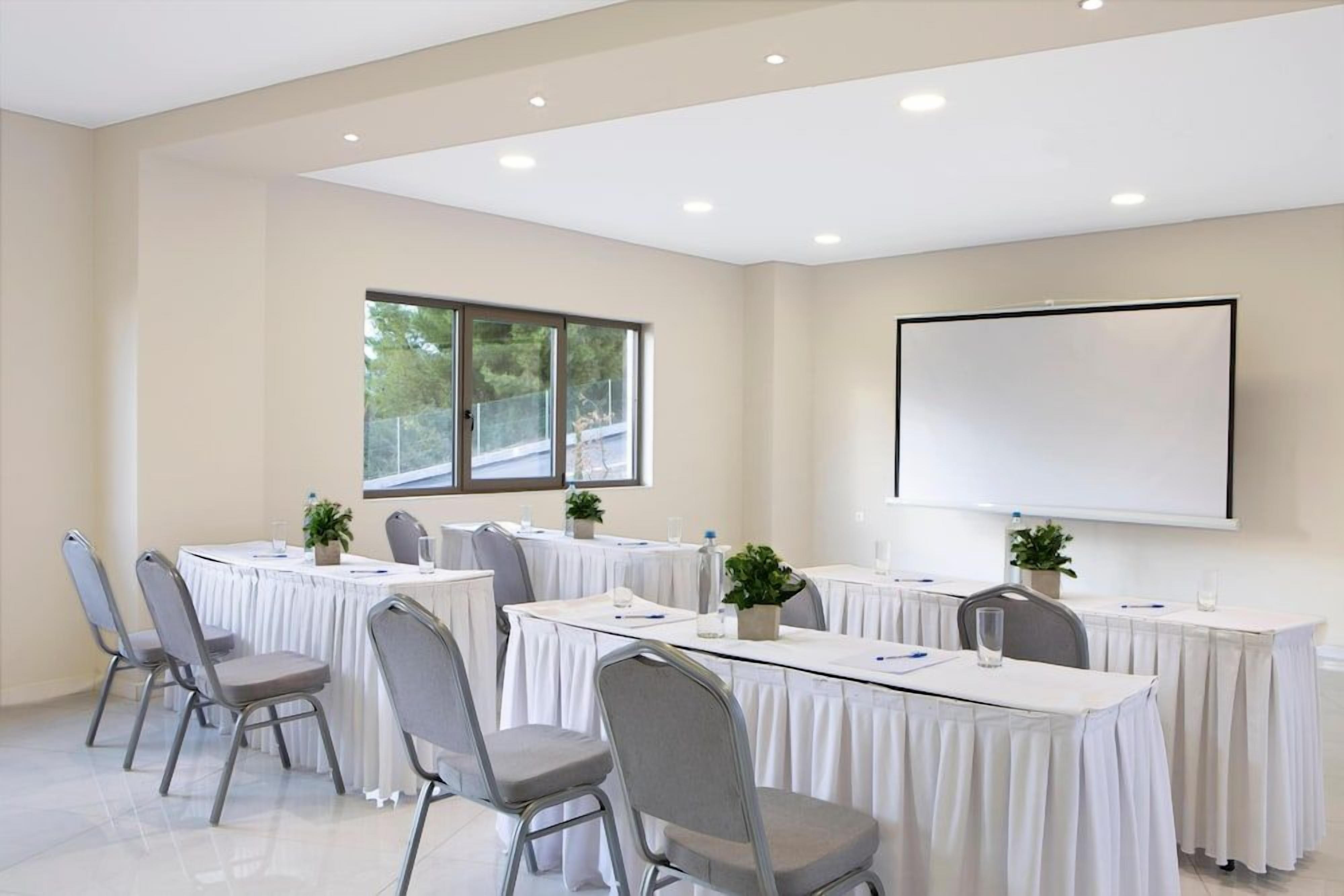 Une salle de conférence équipée d’un grand écran, de plusieurs tables et de chaises, ainsi que de bouteilles d’eau et de stylos.