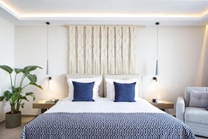 A modern hotel room with a large bed, two bedside tables, a potted plant, and a seating area with a chair.
