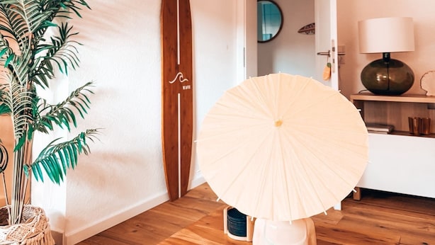 Ein Zimmer mit Holzboden, einer Pflanze, einem Surfbrett, einer Hängelampe und einer Tür zu einem weiteren Raum.