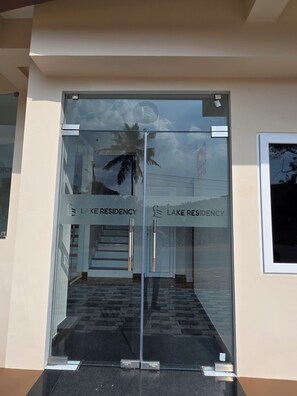 Entrada con puerta de vidrio a un edificio, donde se lee 'LAKE RESIDENCY' en el cristal.