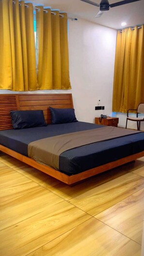 A bedroom with a wooden bed, a nightstand, a chair, and yellow curtains. - Lake Residency (Kadayal)