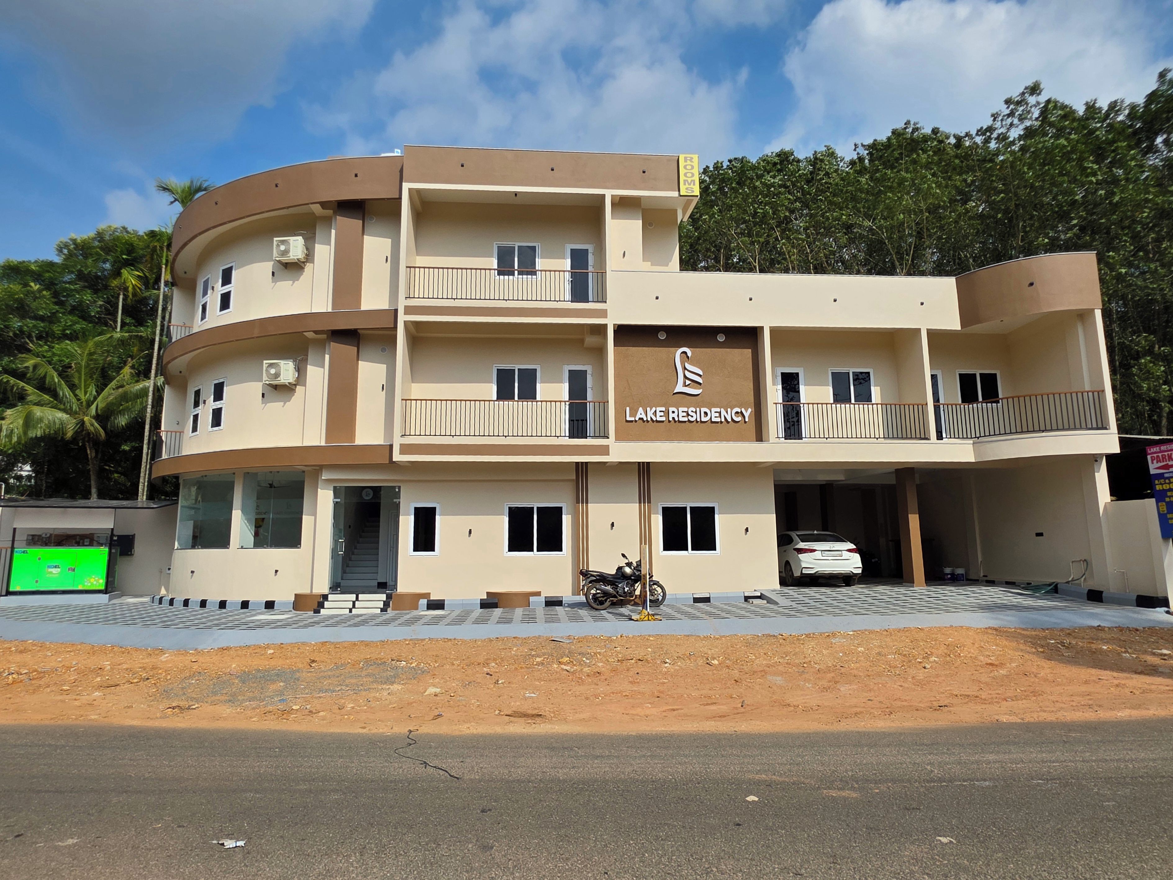 A multi-story building with a sign reading 'Lake Residency.' It has balconies, a car parked in the driveway, and a dirt area in front.