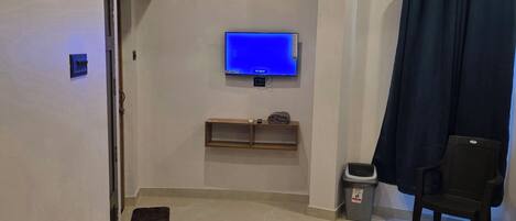 A hotel room with a flat-screen TV mounted on the wall, a ceiling fan, a dark curtain, a trash bin, and a chair.