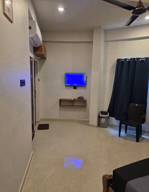 Habitación de hotel con televisor de pantalla plana montado en la pared, ventilador de techo, cortina oscura, cesto de basura y una silla.