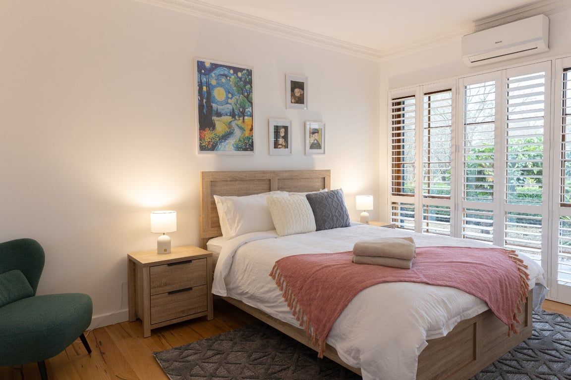 Ein Schlafzimmer mit einem Holz-Kopfteil, einem Bett mit weißer Bettwäsche, einem grünen Sessel, einem Nachttisch mit Lampe und einem Fenster mit Jalousien.
