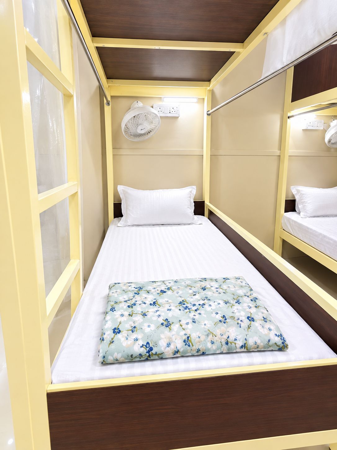 A single bed with a floral bedspread, a bedside fan, and a window with curtains.