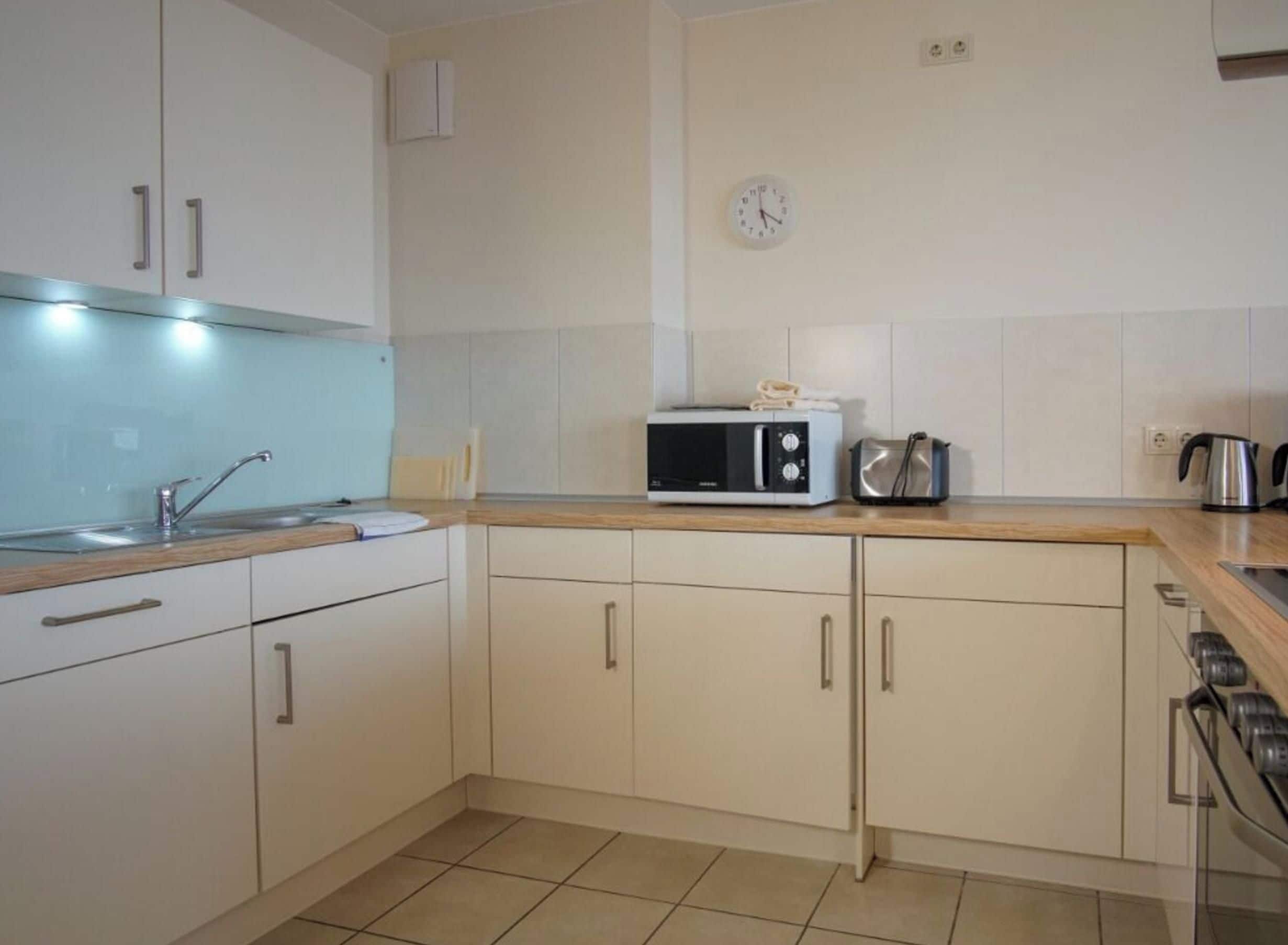 A modern kitchen with white cabinets, a microwave, a kettle, and a clock on the wall.