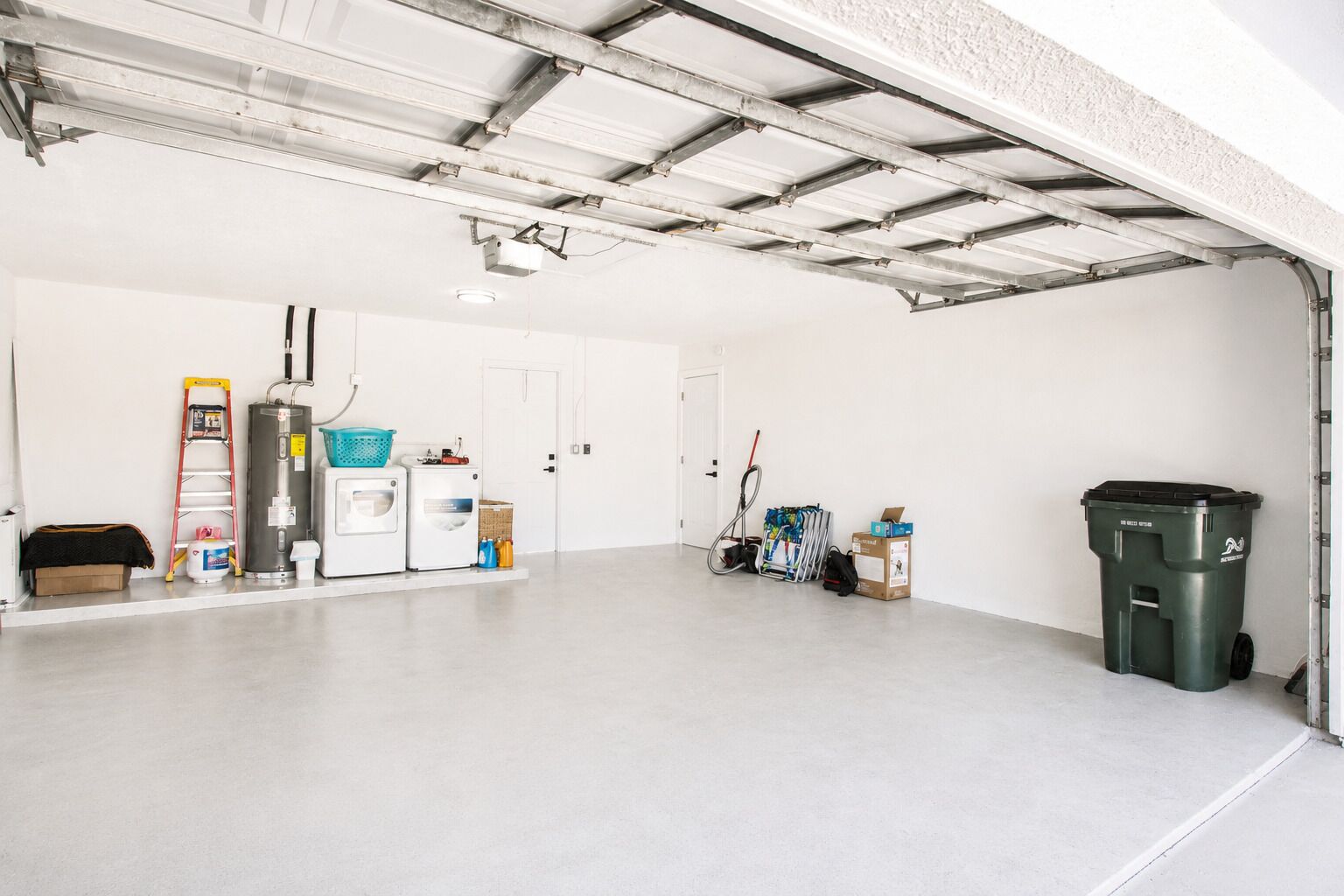 Eine saubere, leere Garage mit einer weißen Tür, einem Warmwasserspeicher, einer Waschmaschine, einer Leiter, Reinigungsmitteln und einem Mülleimer.