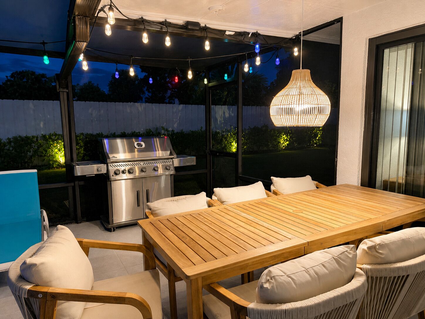An outdoor patio with a wooden table and chairs, a grill, string lights, and a hanging light fixture.