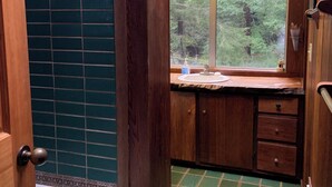 Interior de una cabaña de madera con una bañera, piso de baldosas verdes y ventana con vista a un bosque.
