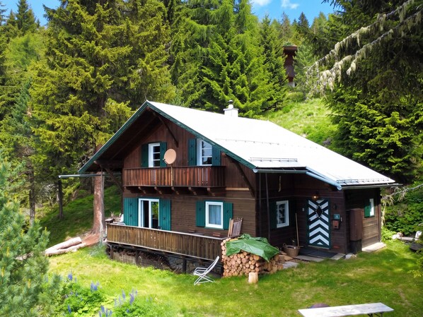 A two-story wooden cabin with a balcony, surrounded by dense forest.