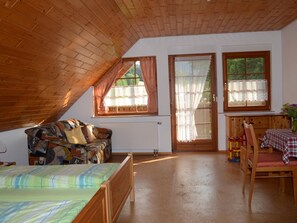 A cozy attic bedroom with a sloped ceiling, a bed, a sofa, a dining table with chairs, and two windows with curtains. - Apartment in the Black Forest with a balcony (Schuttertal)