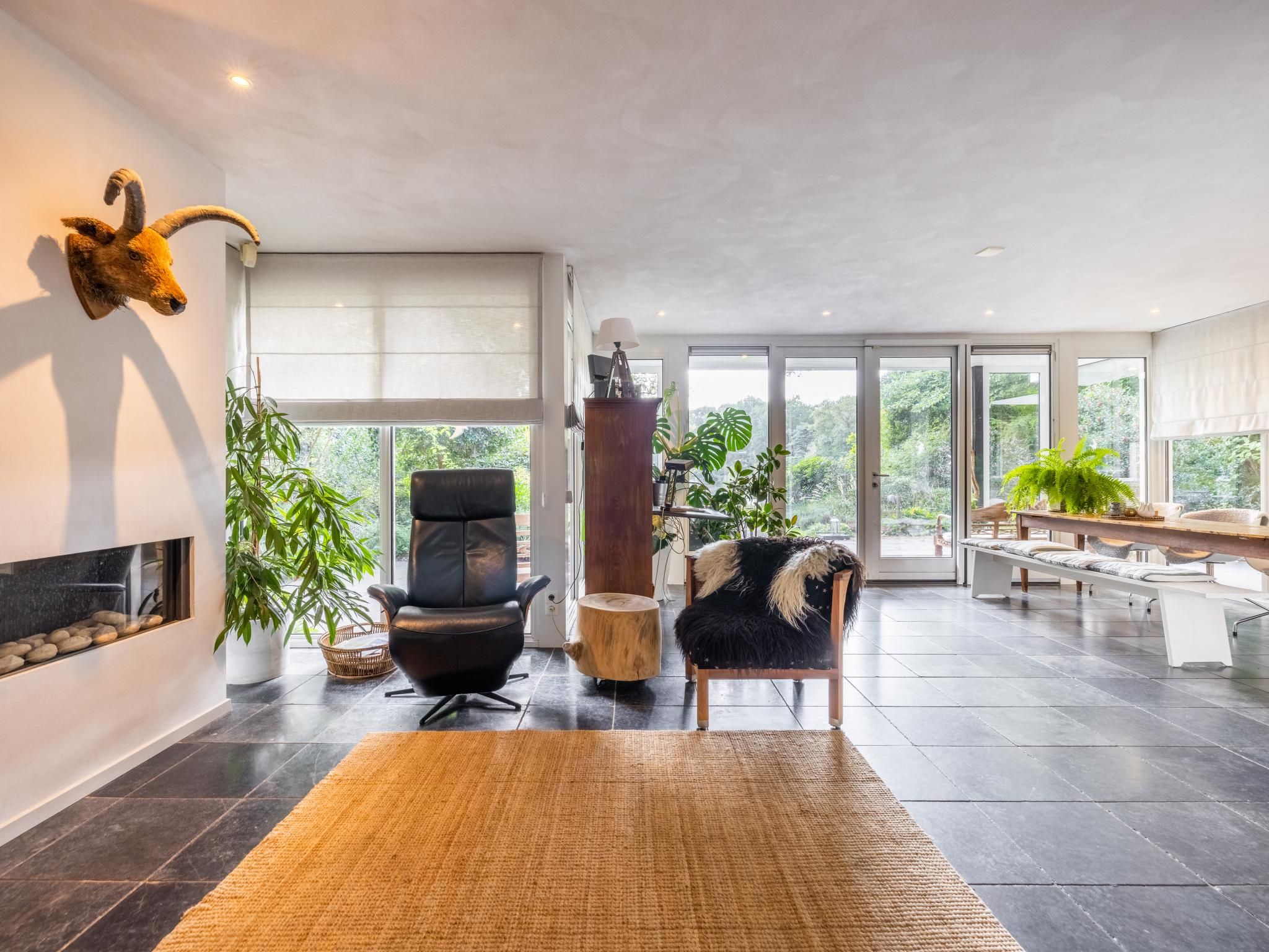 A modern living room with a fireplace, a black leather chair, a wooden side table, and a large window offering a view of greenery.
