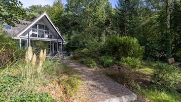 A modern house with a large balcony, surrounded by dense forest and a gravel path.